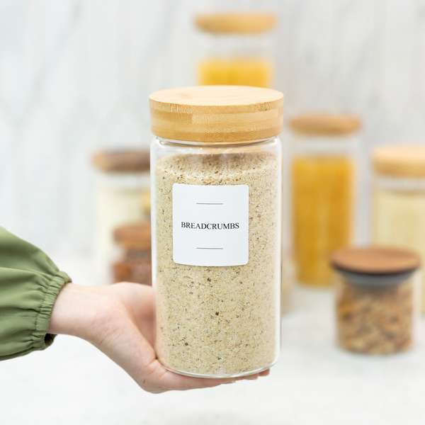 Glass jar labeled 'Breadcrumbs' with a wooden lid held by a hand, with more jars in the background.