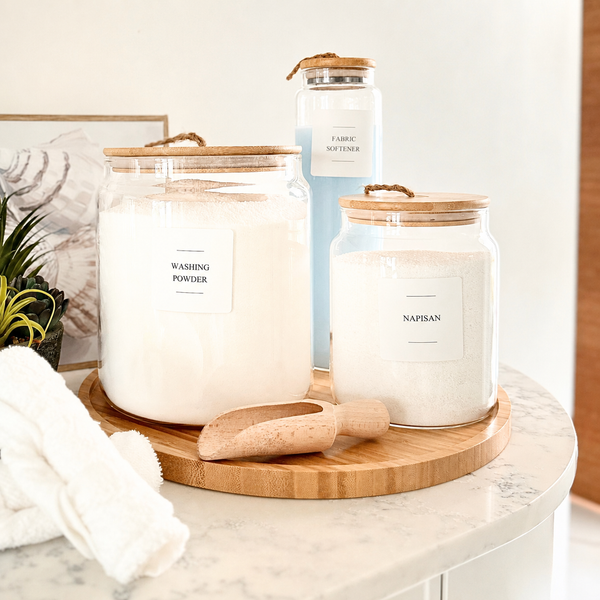 Three clear glass jars with light wood lids and rope handles are arranged on a round wooden tray on a marble countertop. The front-left jar is labeled 'WASHING POWDER' and contains a white powder.