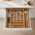 A wooden drawer organizer tray holding a coordinated set of stainless steel cutlery, including knives, forks, spoons, chopsticks, and serving utensils, shown in an open kitchen drawer.