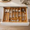 An open kitchen drawer showing a wooden cutlery organizer filled with stainless steel utensils, including spoons, forks, knives, and measuring cups, set in a bright modern kitchen.