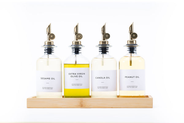 Four round glass oil dispensers with metal spouts on wood tray, ideal for kitchen storage and organisation, featuring sesame oil.