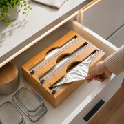 A wooden drawer organizer with sliding aluminum slots holding a foil bag, shown inside an open kitchen drawer with a hand pulling the foil out. Clear glass storage containers and a countertop are visible around it.