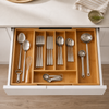 An open kitchen drawer showing a coordinated stainless steel utensil set arranged in wooden divider compartments. Visible items include spoons, forks, knives, a whisk, measuring cups, and serving ladles, set against a white drawer front and light wood floor.