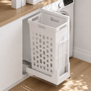 A white under-counter pull-out laundry hamper basket with a metal frame, shown partially extended next to a washing machine in a bright, modern laundry room.