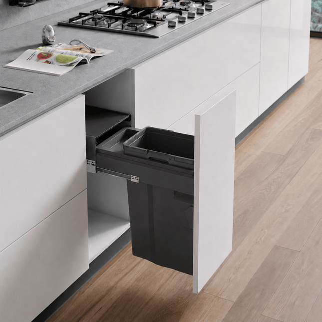 A dark gray under-counter kitchen trash bin with its cabinet door open, set in a modern kitchen with white cabinets, a gray countertop, and light wood flooring.