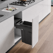 A dark gray under-counter kitchen trash bin with its cabinet door open, set in a modern kitchen with white cabinets, a gray countertop, and light wood flooring.
