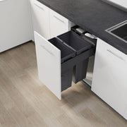 A dark gray under-counter double trash bin with pull-out doors, installed in a white kitchen cabinet next to a dark countertop on a light wood floor.