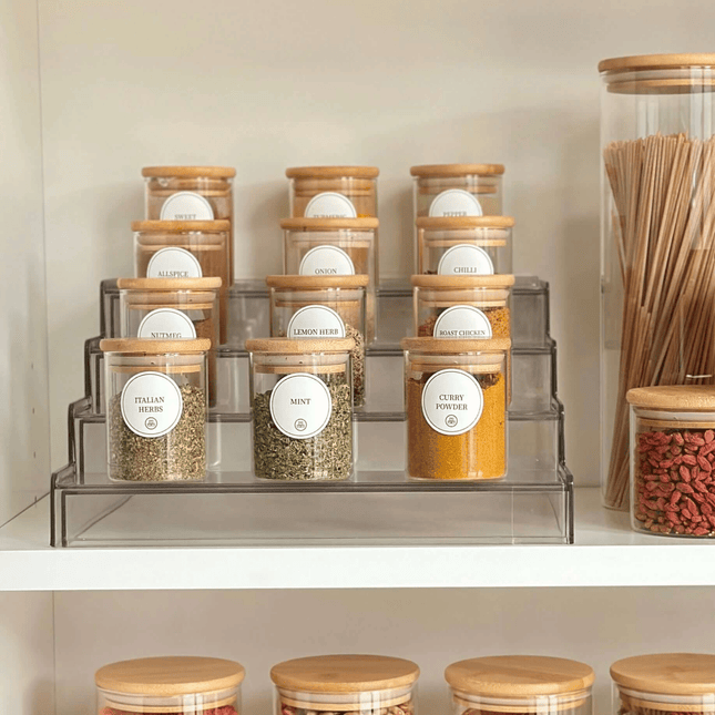 Round glass bamboo herb and spice storage jars organised on a clear tiered spice rack inside a kitchen pantry cupboard