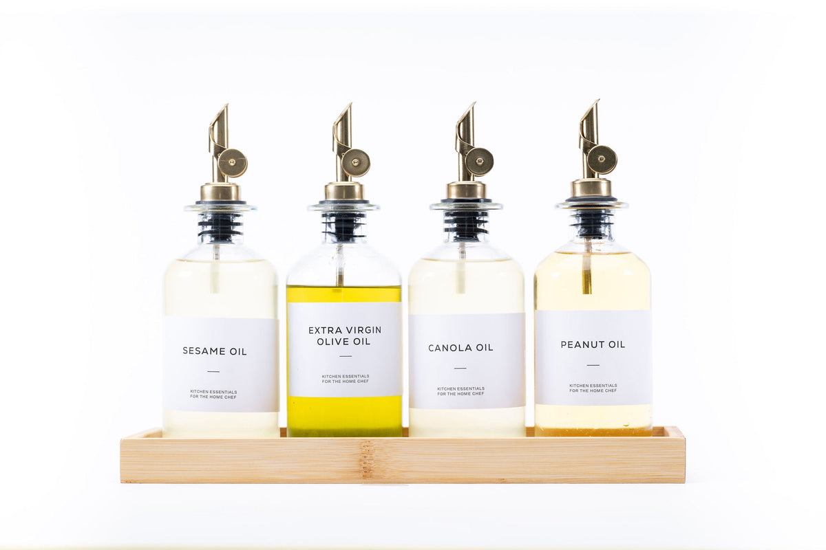 Four round glass oil dispensers with metal spouts on wood tray, ideal for kitchen storage and organisation, featuring sesame oil.