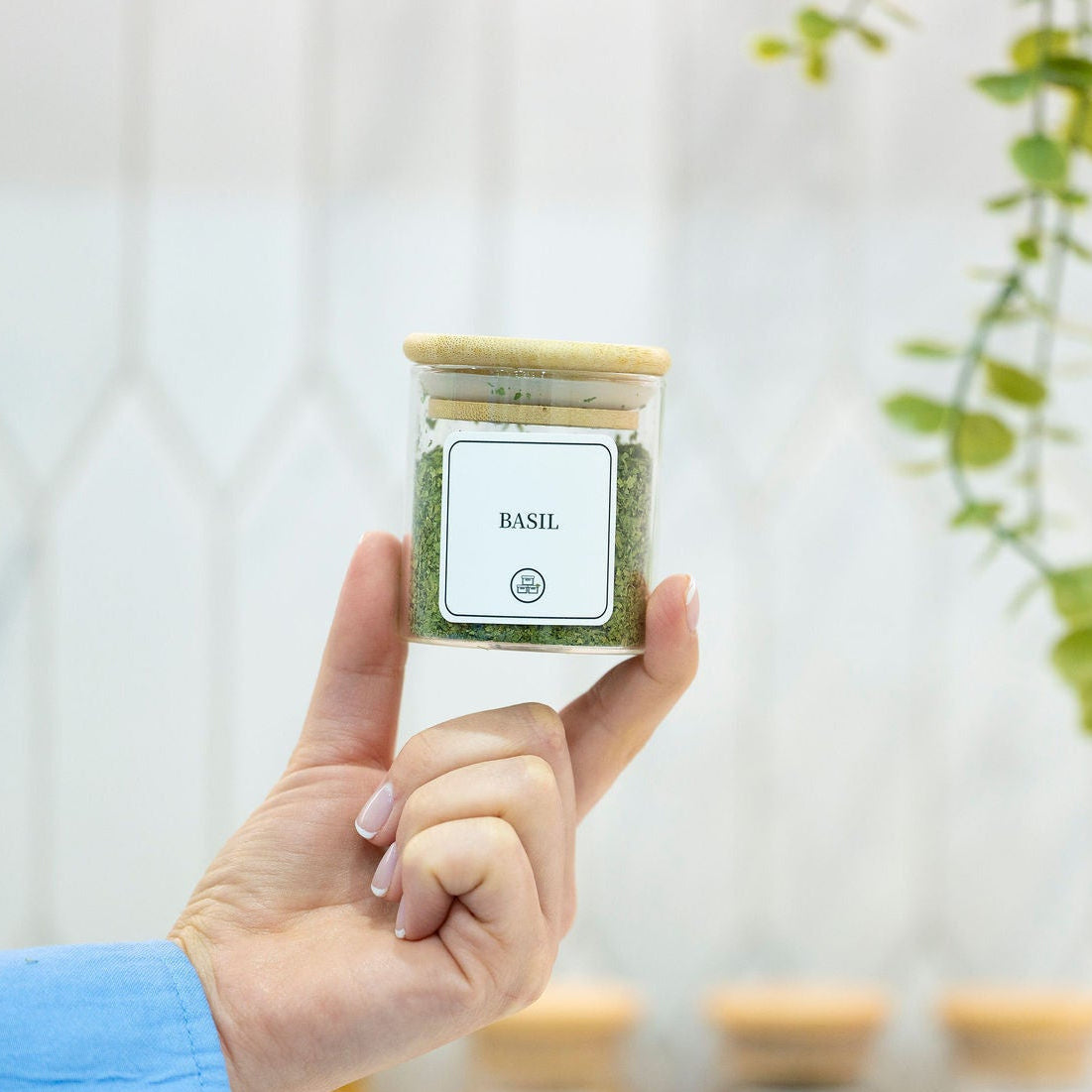 Hand holding a basil-filled Round Glass Spice Jar with Bamboo Lid – 3 Sizes for pantry organisation in a kitchen setting.