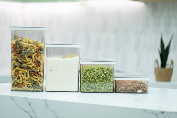 Row of clear airtight pantry containers in different heights holding pasta, rice, peas and almonds on a white marble bench